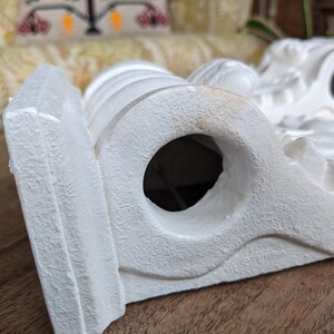May include: White plaster corbel with a decorative design and a round hole in the center. The corbel is a decorative architectural element used to support a shelf or other structure.