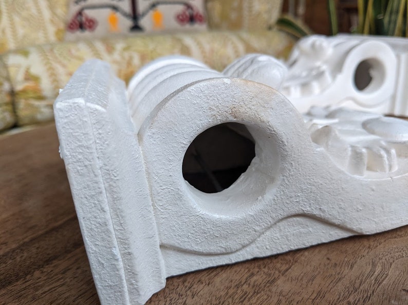 May include: White plaster corbel with a decorative design and a round hole in the center. The corbel is a decorative architectural element used to support a shelf or other structure.