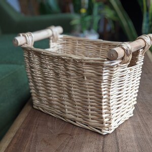 May include: A natural woven wicker basket with two wooden handles. The basket is empty and sitting on a wooden surface.