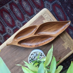 May include: A wooden serving dish shaped like a leaf with four separate compartments. The dish is made of dark brown wood and is sitting on a wooden table.