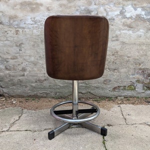May include: A brown leather swivel bar stool with a chrome base and footrest. The stool is in front of a brick and concrete wall.