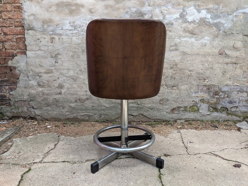 May include: A brown leather swivel bar stool with a chrome base and footrest. The stool is in front of a brick and concrete wall.
