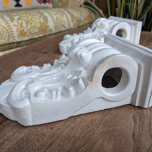 May include: Two white plaster corbels with ornate details and a hole in the center. The corbels are on a wooden surface.