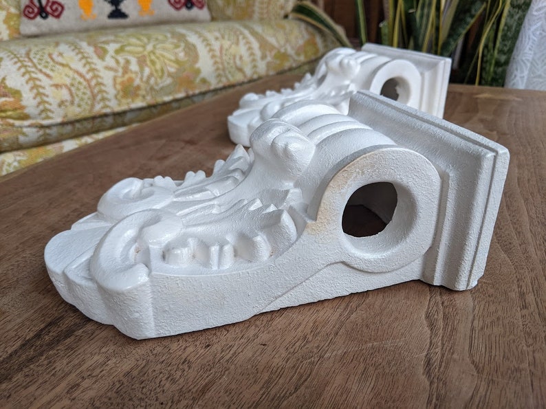 May include: Two white plaster corbels with ornate details and a hole in the center. The corbels are on a wooden surface.