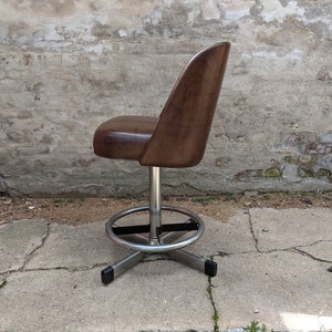 May include: A brown leather swivel bar stool with a chrome base and footrest. The stool is in front of a brick wall.