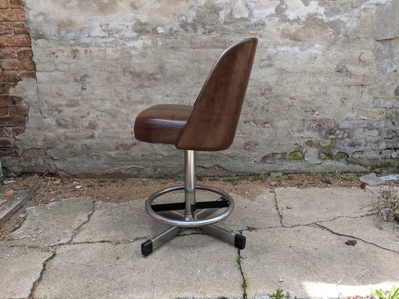 May include: A brown leather swivel bar stool with a chrome base and footrest. The stool is in front of a brick wall.