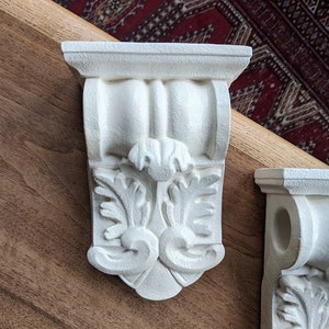 May include: Two white plaster corbels with ornate floral designs. The corbels are mounted on a wooden surface.