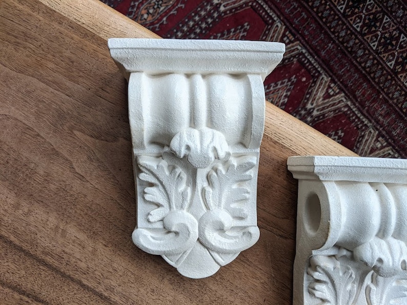 May include: Two white plaster corbels with ornate floral designs. The corbels are mounted on a wooden surface.