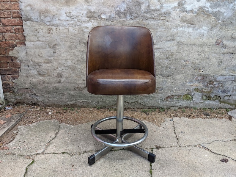 May include: A brown leather swivel bar stool with a chrome base and footrest. The stool has a rounded back and a comfortable seat.
