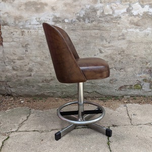 May include: A brown leather swivel bar stool with a chrome base and black footrest. The stool is in a side view and is sitting on a concrete surface.