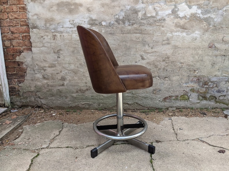 May include: A brown leather swivel bar stool with a chrome base and black footrest. The stool is in a side view and is sitting on a concrete surface.