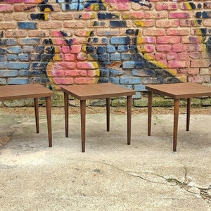 May include: Three brown wooden end tables with square tops and four legs. The tables are arranged in a row against a brick wall with graffiti.