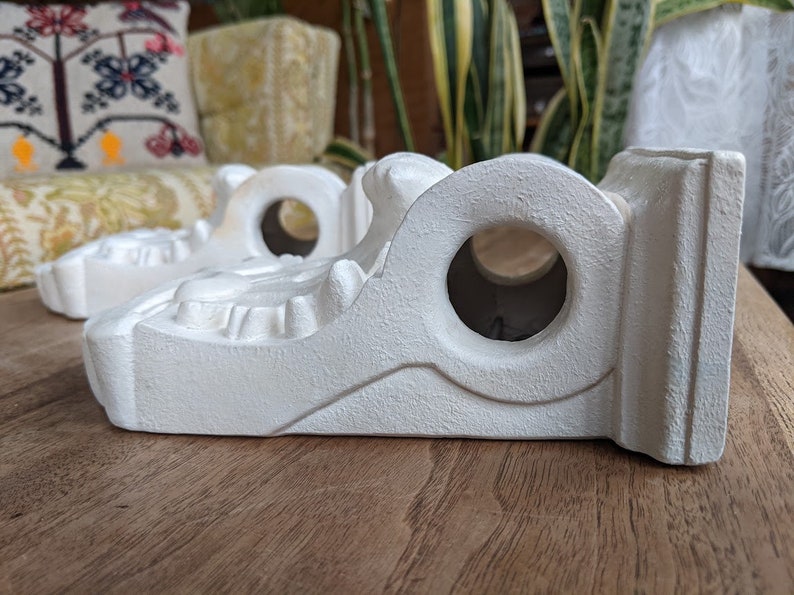May include: Three white ceramic shelf brackets with intricate details and a round hole in the center. The brackets are designed to support shelves or other decorative items.