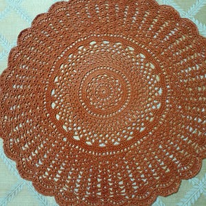 May include: A round, rust-colored crochet doily with a scalloped edge. The doily features a detailed, openwork design with concentric circles radiating from the center. The diameter is approximately 12 inches.