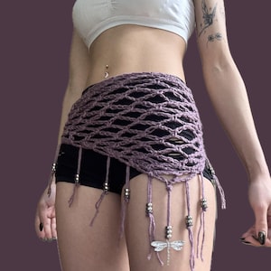 May include: A lavender crochet hip wrap with a net-like design. The wrap features dangling strands with silver beads and a dragonfly charm. The garment is worn over black shorts.