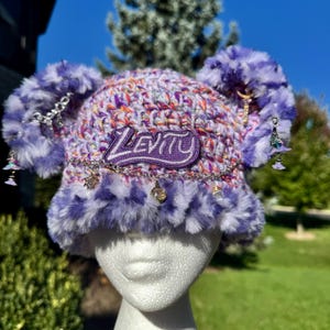 May include: A purple and white variegated crochet beanie featuring two large faux fur pom-poms, a purple patch with the text Levity, and a silver-toned chain draped across the front with small dangling charms and crystals.