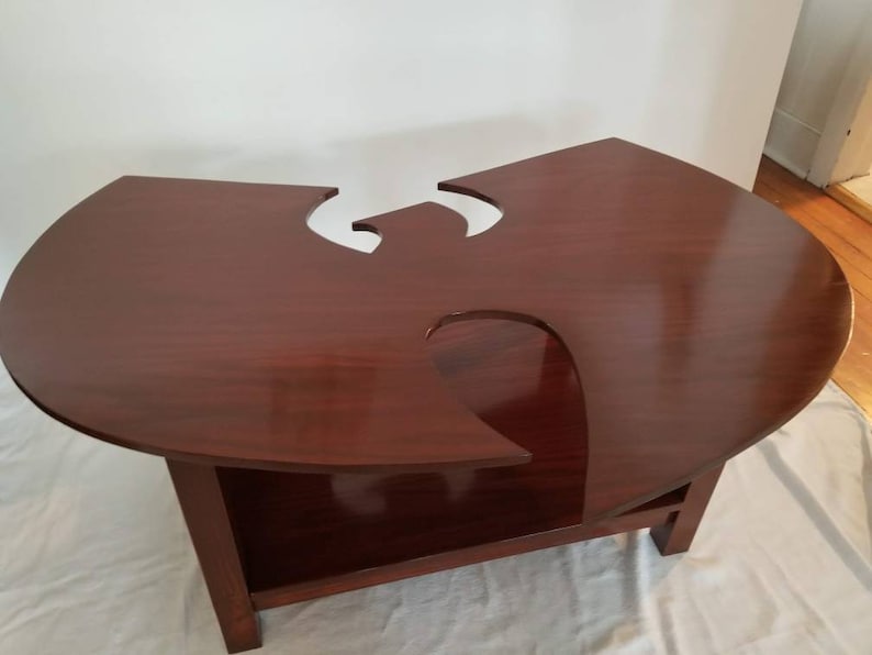May include: A dark brown wooden coffee table with a unique cutout design in the shape of the Wu-Tang Clan logo. The table has a lower shelf for storage.
