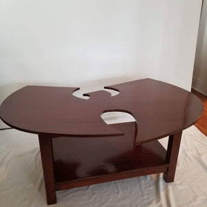 May include: A dark brown wooden coffee table with a unique cutout design in the shape of the Wu-Tang Clan logo. The table has a lower shelf for storage.