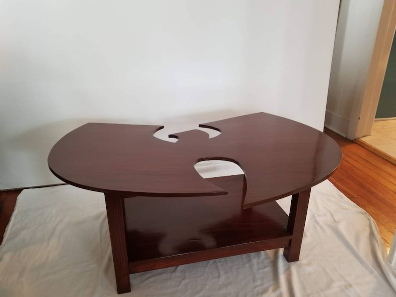 May include: A dark brown wooden coffee table with a unique cutout design in the shape of the Wu-Tang Clan logo. The table has a lower shelf for storage.
