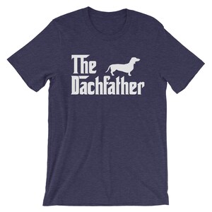May include: Navy blue t-shirt with the text "The Dachfather" in a white, stylized font, accompanied by a white silhouette of a dachshund dog. The shirt is a short-sleeved crew neck.