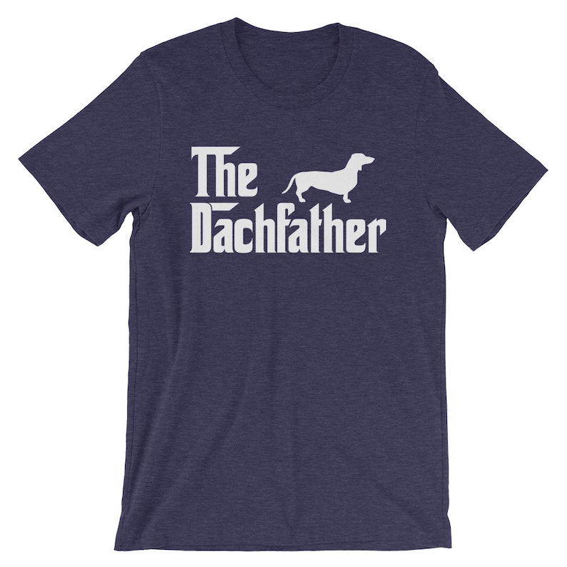 May include: Navy blue t-shirt with the text "The Dachfather" in a white, stylized font, accompanied by a white silhouette of a dachshund dog. The shirt is a short-sleeved crew neck.