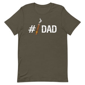 Puede incluir: Una camiseta verde oscuro con un hashtag blanco y la palabra "DAD" impresa en ella. Un cigarro de dibujos animados con humo est&aacute; al lado del hashtag.