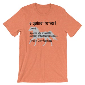 Puede incluir: Una camiseta naranja claro con una silueta blanca de un caballo corriendo. El texto en la camiseta dice "e-quine-tro-vert {noun} Una persona que prefiere la compa&ntilde;&iacute;a de los caballos a la de los humanos. Ver tambi&eacute;n: Crazy horse lady"