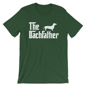 May include: A dark green t-shirt with the text "The Dachfather" in a white, stylized font, accompanied by a white silhouette of a dachshund dog. The shirt is a short-sleeved crew neck.