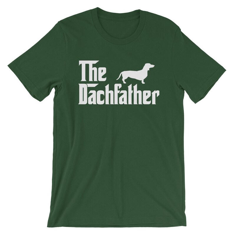 May include: A dark green t-shirt with the text "The Dachfather" in a white, stylized font, accompanied by a white silhouette of a dachshund dog. The shirt is a short-sleeved crew neck.