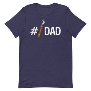 Puede incluir: Una camiseta azul marino con un hashtag blanco y la palabra "DAD" impresa en la parte delantera. Un cigarro de dibujos animados con humo est&aacute; al lado del hashtag.