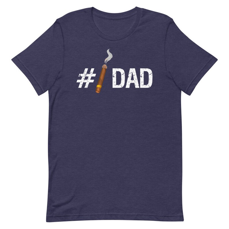 Puede incluir: Una camiseta azul marino con un hashtag blanco y la palabra "DAD" impresa en la parte delantera. Un cigarro de dibujos animados con humo est&aacute; al lado del hashtag.