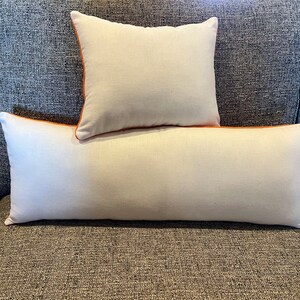 May include: Two decorative pillows, one square and one rectangular, rest on a gray sofa. The pillows are off-white with orange piping around the edges. The sofa has a textured, gray fabric.