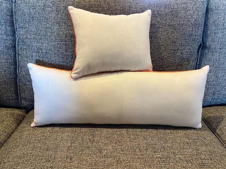 May include: Two decorative pillows, one square and one rectangular, rest on a gray sofa. The pillows are off-white with orange piping around the edges. The sofa has a textured, gray fabric.
