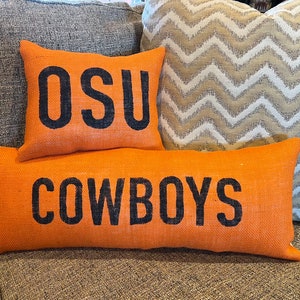 May include: Orange burlap pillows with black text. One pillow reads "OSU" and another reads "COWBOYS". A patterned pillow is in the background. The pillows are on a gray couch.