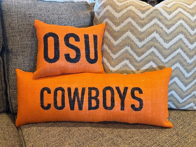 May include: Orange burlap pillows with black text. One pillow reads "OSU" and another reads "COWBOYS". A patterned pillow is in the background. The pillows are on a gray couch.