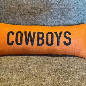 May include: Orange rectangular pillow with the word "COWBOYS" printed in black. The pillow is made of a textured, burlap-like material and rests on a gray upholstered surface. A decorative accent for a home.