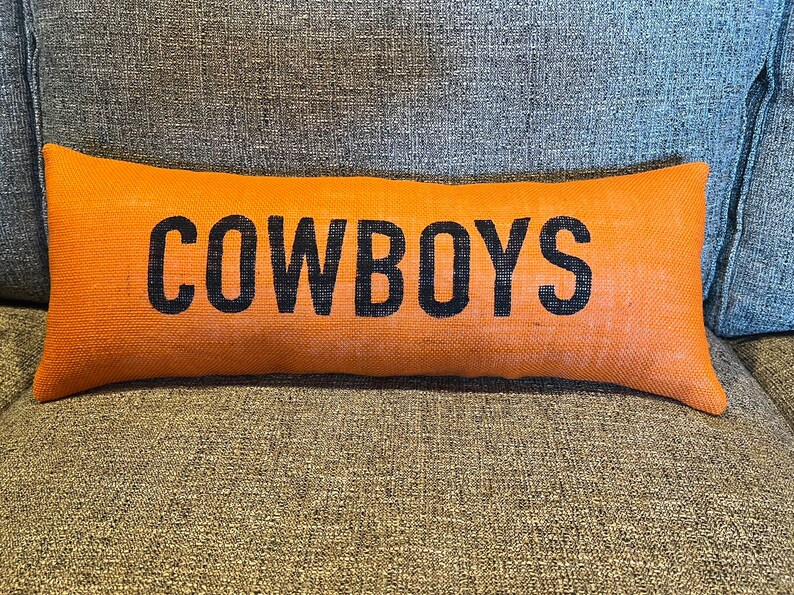 May include: Orange rectangular pillow with the word "COWBOYS" printed in black. The pillow is made of a textured, burlap-like material and rests on a gray upholstered surface. A decorative accent for a home.