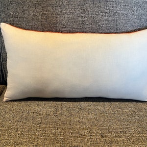 K&ouml;nnte beinhalten: Ein wei&szlig;es rechteckiges Kissen mit orangefarbenem Rand, das auf einem grauen Sofa liegt.