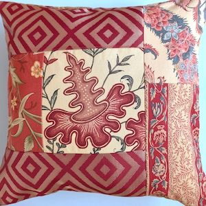 May include: A square throw pillow featuring a patchwork design of various fabrics. The pattern includes sections of red and tan geometric diamonds, a central panel with a large red organic floral motif on cream, and side panels with floral prints.