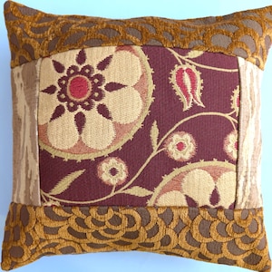 May include: A square throw pillow featuring a central panel with a deep burgundy background, adorned with beige and red floral patterns. The pillow is framed by textured, golden-brown scalloped borders and vertical side panels in a light beige tone.