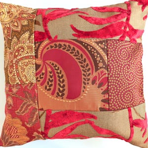 May include: A square throw pillow featuring a patchwork design of various fabrics in shades of red, brown, and tan. The surface includes panels with paisley patterns, solid red velvet leaf motifs, and a textured section with small circular dots.