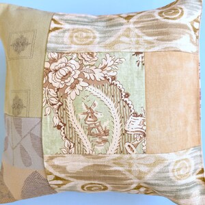 May include: Square cushion with a patchwork design. The cushion features various fabric patterns in shades of beige, tan, and green. One section displays a floral and windmill motif. The cushion is likely used for home decor.