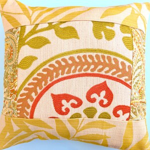 May include: A square throw pillow featuring a central panel with an orange and olive green embroidered circular motif on a cream background, framed by borders of yellow leaf patterns and side panels with an intricate orange and green paisley print.
