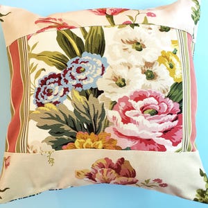May include: A square throw cushion featuring a central panel with a vibrant floral print of pink, blue, yellow, and white flowers on a cream background, framed by vertical striped borders in shades of red, green, and cream with floral accents.