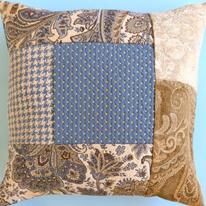 May include: Square patchwork cushion with a variety of patterned fabrics. The cushion features a central blue square with yellow dots, surrounded by sections of paisley, houndstooth, and textured beige fabrics. A decorative home accent.