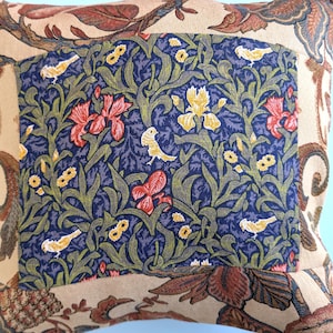 May include: A square throw pillow featuring a central rectangular panel with a dark blue background, green foliage, yellow birds, and red and yellow flowers. The surrounding border is a light tan fabric with a brown and green floral pattern.