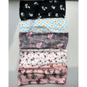 May include: Five different fabric headbands with various patterns. The top headband is black with white flowers. The second headband is light blue with white gingham and white flowers. The third headband is gray with a black and white print of books and the text "Keep Calm and Read On". The fourth headband is white with small red and green flowers. The bottom headband is pink with small red, green, and yellow flowers.
