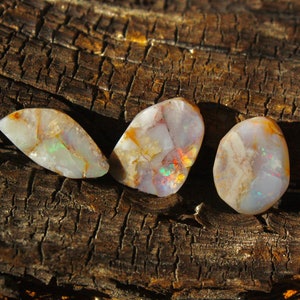 May include: Three iridescent opal stones with a white, grey, and brown colour scheme. The stones are sitting on a piece of weathered wood.