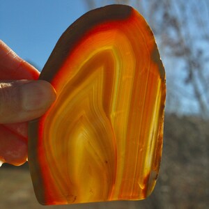 May include: A translucent agate stone slice, showcasing bands of orange, yellow, and brown hues. The natural gemstone is held up against a blue sky, revealing its layered structure and unique patterns. The stone's edges are rough and natural.