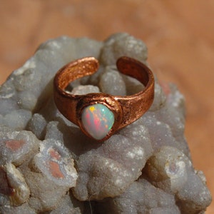 May include: A copper ring with a white opal gemstone set in a bezel setting. The ring is on a white and gray rock background.